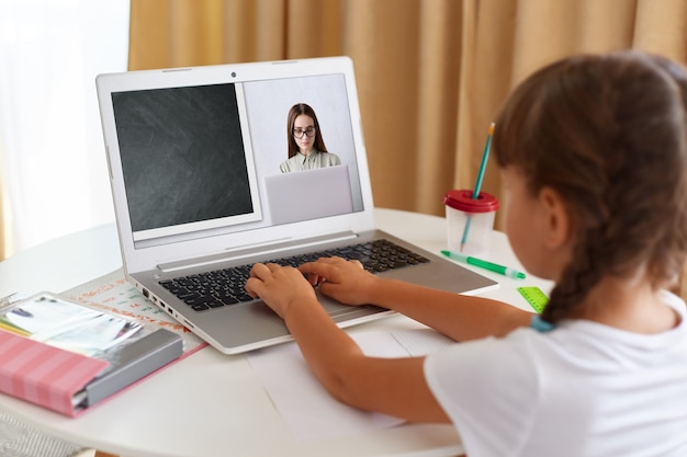 Retrato de vista posterior de colegiala con camiseta blanca sentada frente a la computadora portátil y mirando la pantalla, con lección en línea a través de cámara web, educación a distancia.