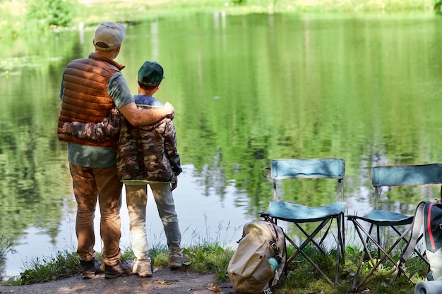Retrato de vista posterior de cuerpo entero de padre e hijo de pie junto al lago y disfrutando de la naturaleza durante el senderismo o el viaje de pesca, espacio de copia