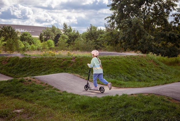 Retrato de vista posterior de una linda colegiala caucásica con casco que disfruta divirtiéndose montando scooter en el parque asfaltado de la calle trackxAin al aire libre en un día soleado Actividades deportivas saludables para niños al aire libre