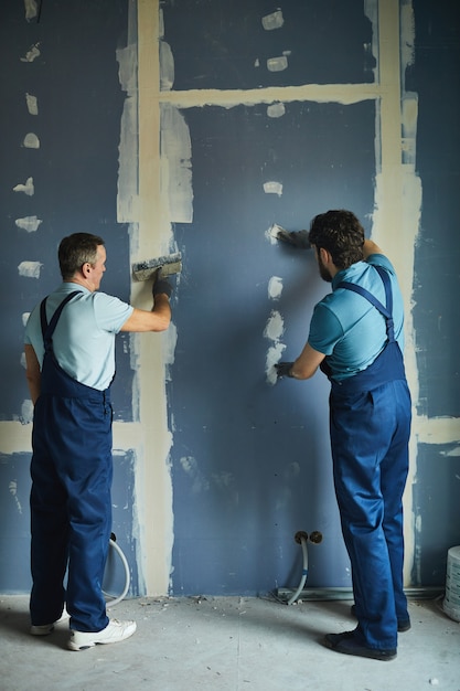 Retrato de vista posterior de longitud completa de dos trabajadores de la construcción que construyen paredes de yeso mientras se renueva la casa, espacio de copia