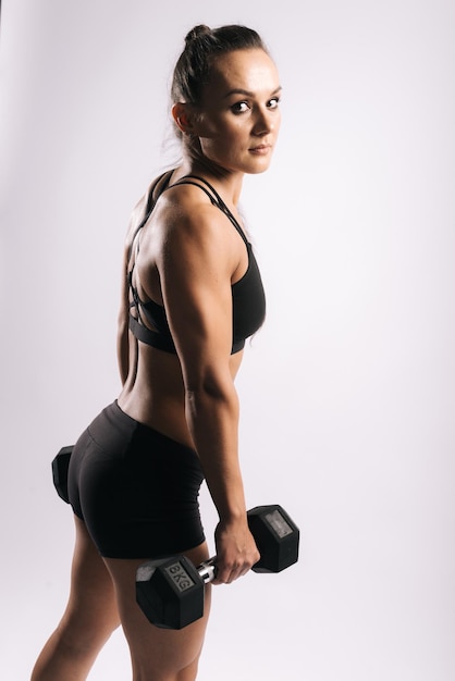 Retrato de vista posterior de mujer joven sexy muscular en ropa deportiva negra. Trabajando con una mancuerna pesada sobre fondo blanco aislado. Chica musculosa deportiva mirando a la cámara