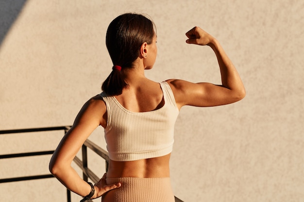 Retrato de vista posterior de niña aturdida con cabello oscuro y cola de caballo con top deportivo de pie con la mano levantada, mostrando sus bíceps y tríceps, resultados del entrenamiento diario.