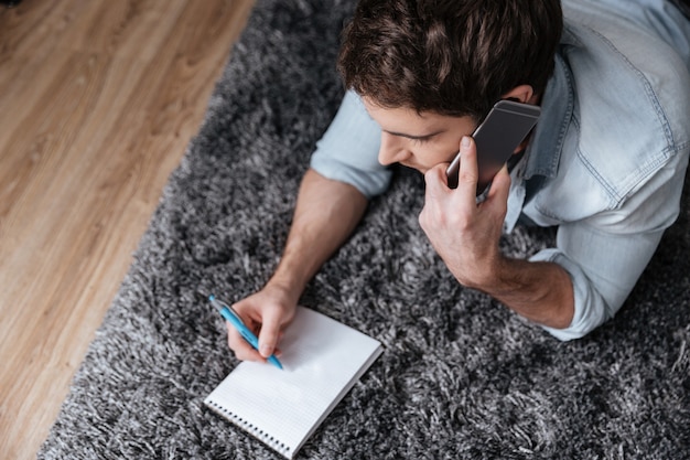 Retrato de vista superior de un hombre casual escribiendo en el bloc de notas y hablando por teléfono móvil en la alfombra en casa