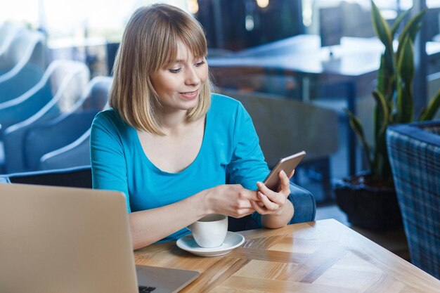 Retrato de vista superior de joven mujer rubia feliz en camiseta azul, sentada en la cafetería y sosteniendo su teléfono móvil inteligente y mensajes de texto. tiro del estudio de interior.