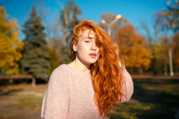 Retratos de uma encantadora garota ruiva com um rosto bonito. Garota posando no parque outono em um suéter e uma saia de cor coral. A menina tem um humor maravilhoso