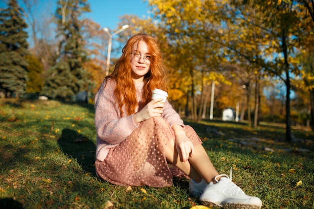 Retratos de uma encantadora garota ruiva de óculos e rosto bonito. Garota posando no parque outono em um suéter e uma saia de cor coral.