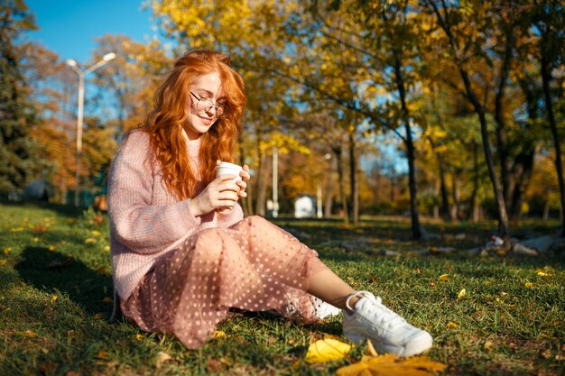 Retratos de una encantadora pelirroja con gafas y una cara bonita. Chica posando en el parque otoño en un suéter y una falda de color coral.