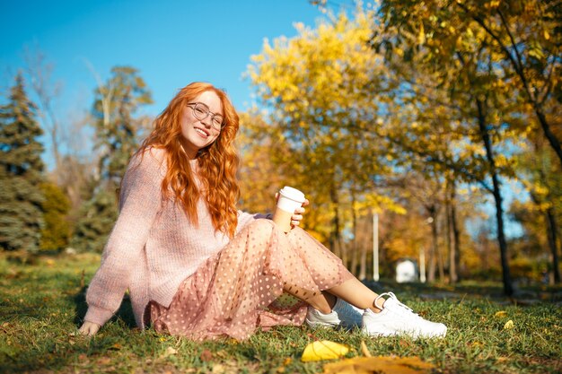 Retratos de una encantadora pelirroja con gafas y una cara bonita. Chica posando en el parque otoño en un suéter y una falda de color coral.