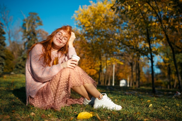 Retratos de una encantadora pelirroja con gafas y una cara bonita. Chica posando en el parque otoño en un suéter y una falda de color coral.