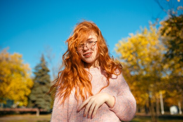 Retratos de una encantadora pelirroja con una linda cara. Chica posando en el parque otoño en un suéter y una falda de color coral. La chica tiene un humor maravilloso