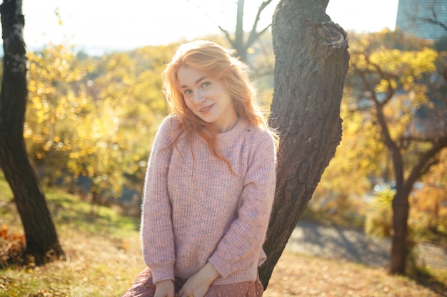 Retratos de una encantadora pelirroja con una linda cara. Chica posando en el parque otoño en un suéter y una falda de color coral. En manos de una niña una hoja amarilla