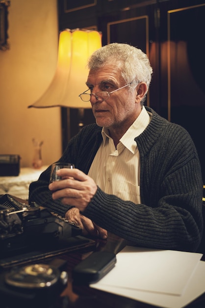 Foto retro-senior-schriftsteller mit brille, der am schreibtisch neben einer veralteten schreibmaschine sitzt, mit einem glas whiskey und nachdenkt.