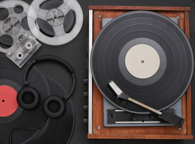 Retro-Vinylplattenspieler mit Schallplatten, magnetischer Audiospule, Audiokassette und Stereokopfhörern auf schwarzem Hintergrund. Ansicht von oben. Flach legen