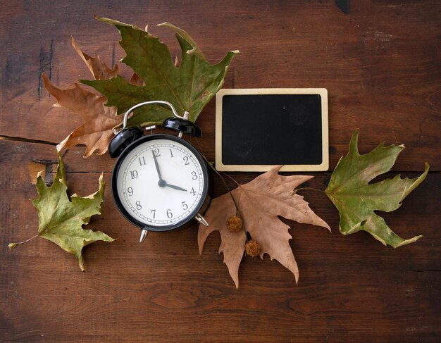 Retroceso a la hora de verano Reloj negro y hojas de otoño en madera
