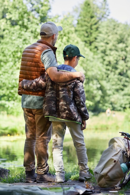 Retrovisor de corpo inteiro de pai e filho à beira do lago, curtindo a natureza durante uma caminhada ou pescaria