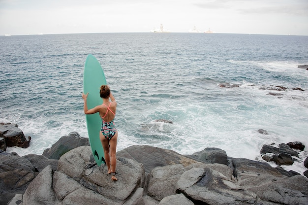 Retrovisor garota sexy no maiô multicolor em pé com as ondas na praia rochosa do oceano Atlântico olhando para a distância