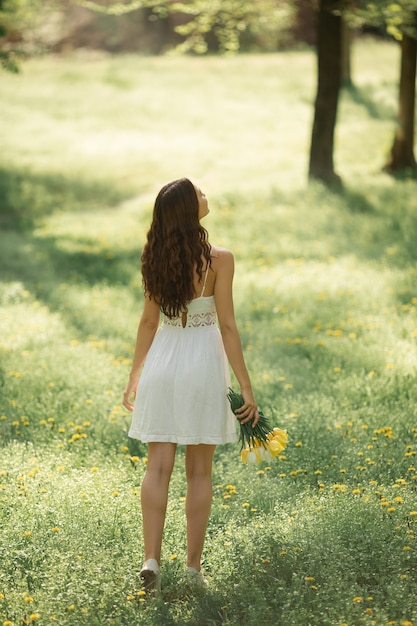 Retrovisor og mulher atraente em um vestido branco com um buquê de flores da primavera caminhando contra