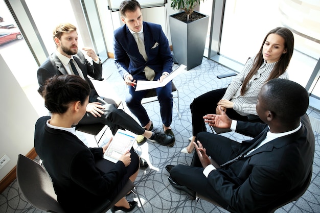 Reunião matinal. Grupo de cinco jovens discutindo algo enquanto estão sentados à mesa no escritório juntos