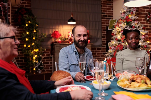 Reunión familiar en la mesa de la cena de Navidad, amigos bebiendo vino espumoso, celebrando la Navidad juntos. Hombre sonriente sosteniendo un vaso, feliz gente diversa comiendo platos tradicionales en la fiesta de casa