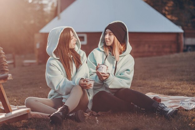 Reuniones en el patio de una pequeña casa de madera. Dos lindas chicas con sudaderas azules idénticas están sentadas en una manta con tazas en las manos y mirándose.