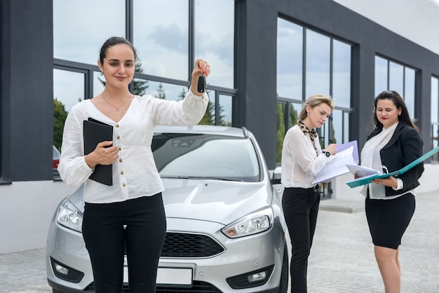Revendedor segurando as chaves do carro novo. Três senhoras com pastas posando do lado de fora do showroom
