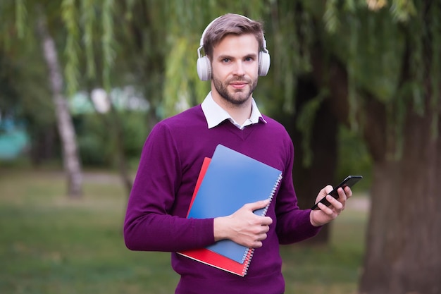 Revise su correo electrónico, escuche música con auriculares y sostenga el cuaderno, chico guapo con auriculares, relájese en el parque, use el teléfono, vida moderna, educación, tecnología, estudiante, obtenga información del estudio de libros en línea.