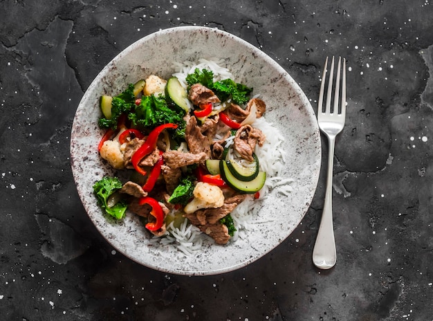 Revuelva la carne de res con calabacín, coliflor, pimiento dulce, col rizada y arroz sobre un fondo oscuro, vista superior, comida de estilo asiático