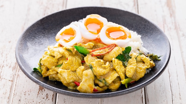 Revuelva el pollo frito con pasta de curry verde, arroz al vapor y huevo cocido con yema líquida en un plato de cerámica negra sobre fondo blanco de textura de madera vieja, Pud Keow Wan Gai, comida tailandesa