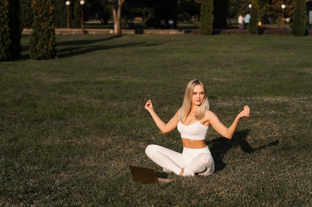 Rezar y meditar en posición de loto en línea con una laptop Fit girl está sentada y meditando en el césped del parque