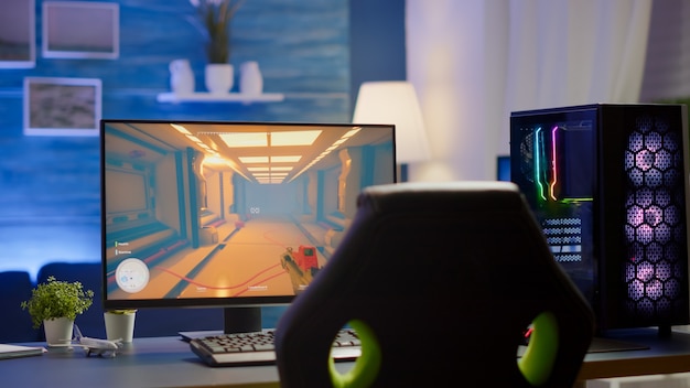 RGB leistungsstarkes PC-Gamer-Rig mit Ego-Shooter-Spiel auf dem Bildschirm. Monitor steht auf dem Tisch im Heimspielraum. Gemütliches Zimmer mit modernem Design wird mit warmem Neonlicht beleuchtet.