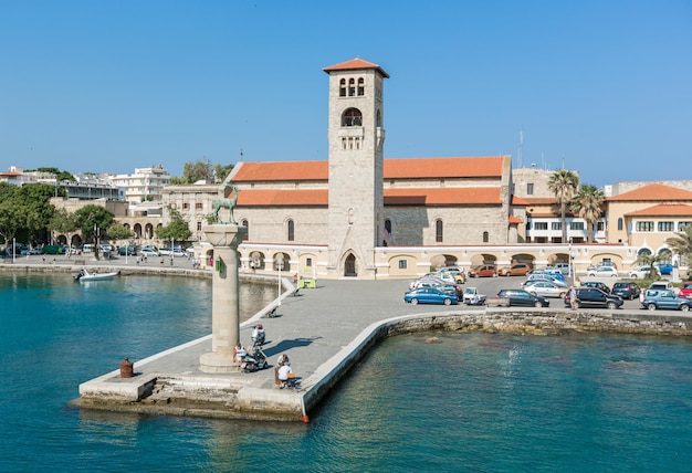 RHODES GRÉCIA 12 de junho de 2015 Vista em um porto e capela do porto de Mandaki Pessoas andando em uma calçadaxA observando estátuas de veados e Forte de São Nicolau com o farol da costa em Rodes Grécia