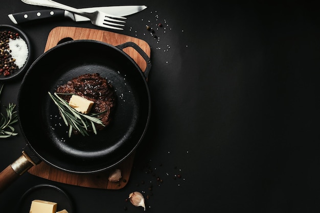 Ribeye cocido bife tabla de sartén de hierro con sal ajo cuchillo horquilla mantequilla fondo negro