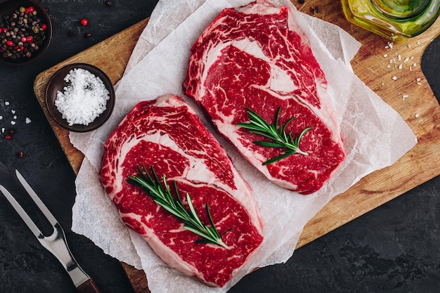 Ribeye Steak Carne de res fresca cruda con sal y romero lista para la barbacoa sobre fondo de piedra oscura