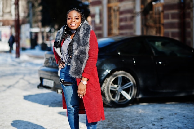 Rica mujer afroamericana en abrigo rojo y pieles en la calle de invierno en un clima soleado contra el coche negro de negocios. Mujer negra con estilo.