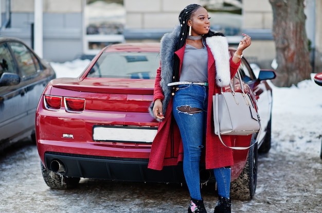 Rica mujer afroamericana en abrigo rojo y pieles contra muscle car rojo. Negro elegante exitosa mujer de negocios.