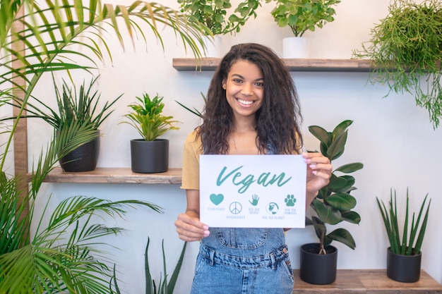 Richtige Wahl. Ziemlich schlanke junge Frau mit Bezeichnung Plakat und Schriftzug vegan stehend lächelnd