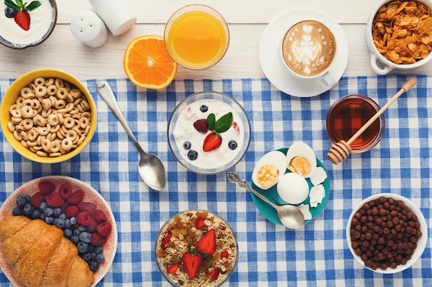 Rico fondo del menú de desayuno. Croissants crujientes franceses, muesli, vaso de naranja fresca, yogur, café, bayas, miel y huevos cocidos para comidas sabrosas por la mañana en manteles a cuadros, vista superior