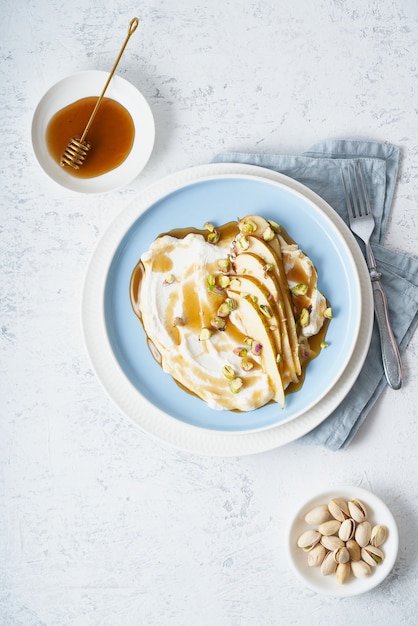 Ricotta con peras, pistachos y miel o jarabe de arce en placa azul sobre mesa blanca.