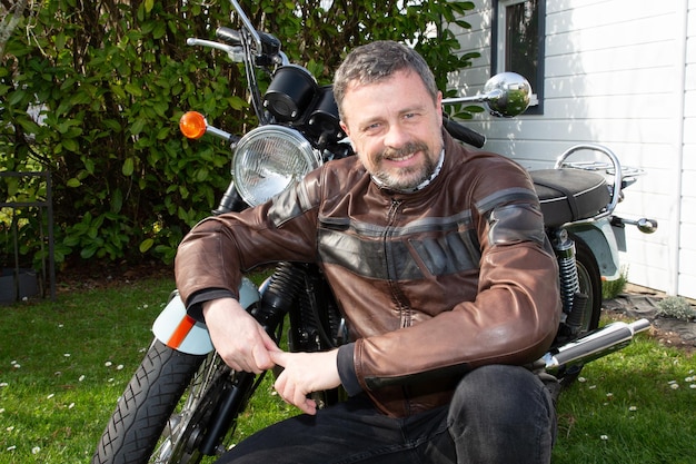 Rider hombre en cuero marrón moto chaqueta de moda sentarse al lado de la motocicleta retro vintage al aire libre