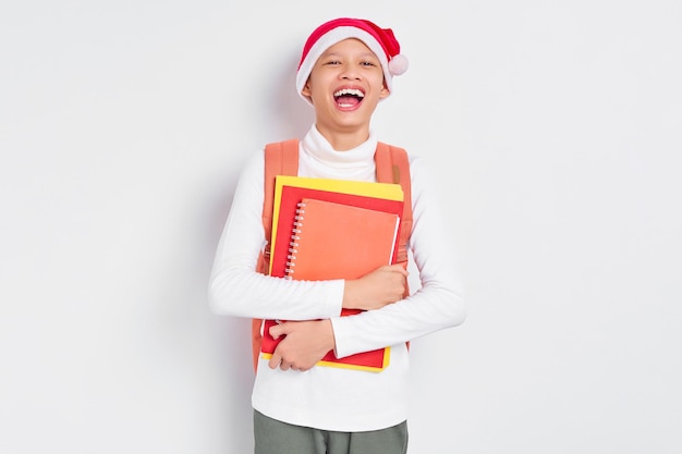 Ríe y es divertido y apuesto joven estudiante asiático con un sombrero de Navidad usando una camiseta con mochila sosteniendo un libro aislado de fondo blanco