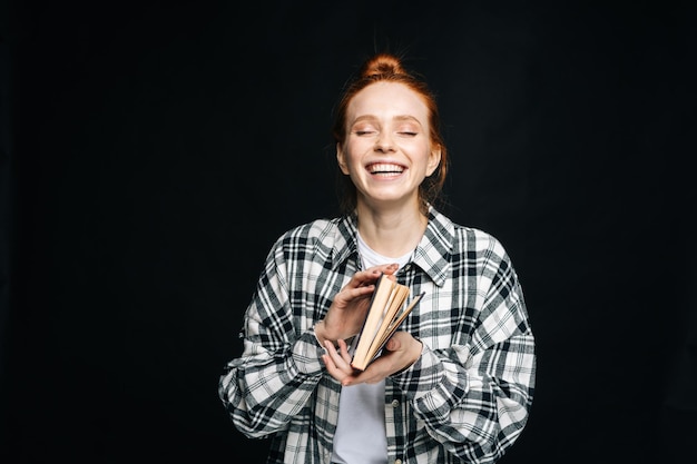 Riendo alegre joven estudiante de pie sosteniendo un libro abierto sobre fondo negro aislado