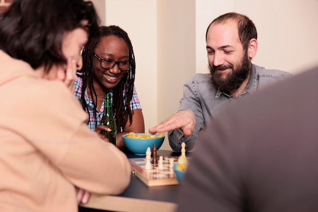 Riendo a amigos cercanos sentados a la mesa en la sala de estar mientras juegan al ajedrez. Amigos inteligentes multiétnicos que disfrutan de juegos de mesa de estrategia mientras se divierten juntos en casa.