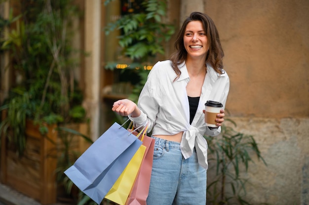 Riendo bonita adicta a las compras femenina milenaria europea con muchas bolsas de compras y una taza de café para llevar