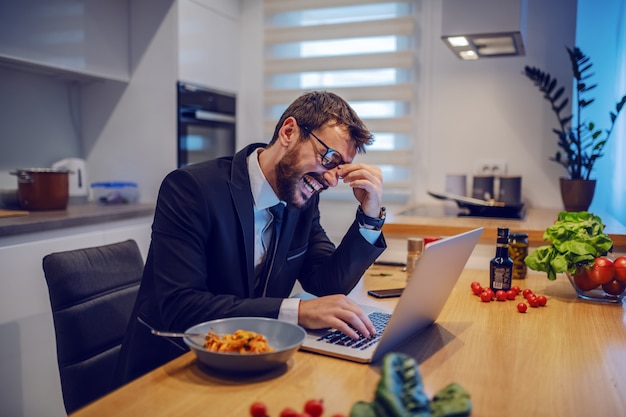 Riendo caucásico barbudo hombre de negocios en traje sentado en la mesa de comedor y usando la computadora portátil. Al lado del portátil hay un plato con pasta.