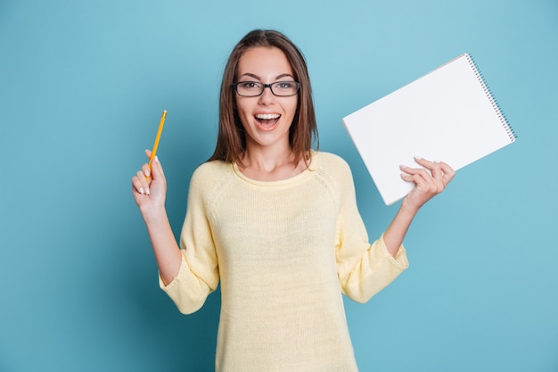 Riendo a chica guapa alegre sosteniendo el cuaderno aislado en el fondo azul.