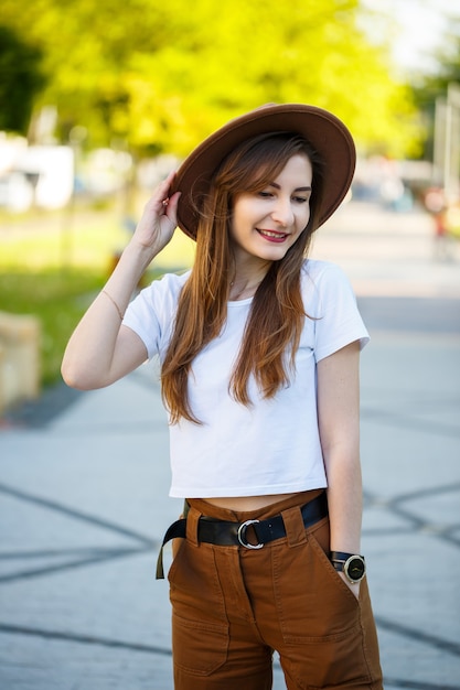 Riendo a chica morena de piel pálida con ropa de moda de pie en un parque disfrutando de una mañana soleada. Retrato de una señorita exquisita con un sombrero y una camiseta blanca posando cerca de un callejón del parque.