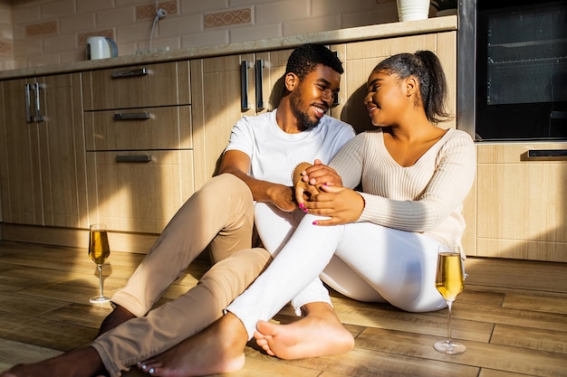 Riendo joven pareja étnica afro saliendo con champán en casa mientras está sentado en el suelo cálido