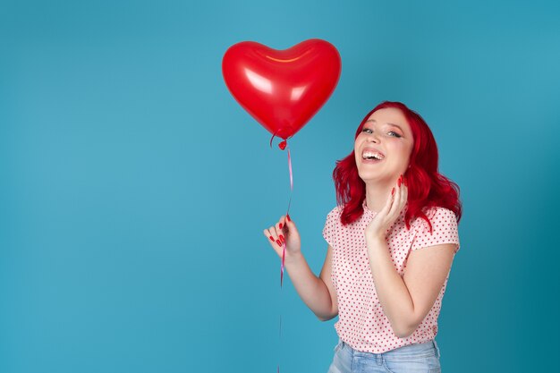 Riendo a mujer joven con cabello rojo sosteniendo un globo volador rojo en forma de corazón