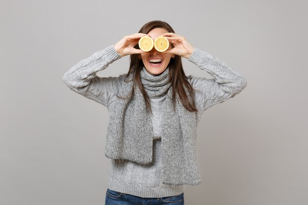 Riendo a mujer joven en suéter gris, bufanda que cubre los ojos con limones aislados sobre fondo de pared gris. Estilo de vida de moda saludable, personas emociones sinceras, concepto de estación fría. Simulacros de espacio de copia.