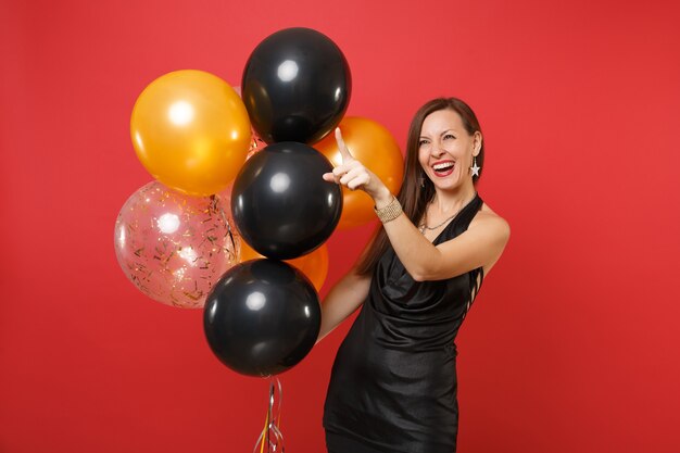 Riendo a mujer joven en vestido negro celebrando señalar con el dedo índice sosteniendo globos de aire aislados sobre fondo rojo brillante. Día de San Valentín, concepto de fiesta de vacaciones de maqueta de cumpleaños de feliz año nuevo.